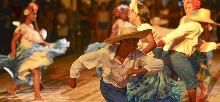 #paratodosverem: A fotografia mostra um grupo de pessoas dançando carimbó. O primeiro plano mostra um rapaz usando chapéu de palha, calça azul e blusa branca com babados, fazendo uma passada da dança, da esquerda para a direita. Ao fundo, mulheres vestindo saias azuis floridas, blusas brancas de babados e tiaras de flores rosa nos cabelos dançam com rapazes vestidos da mesma forma que o primeiro. No fundo da imagem, é possível ver o público que assiste à apresentação.